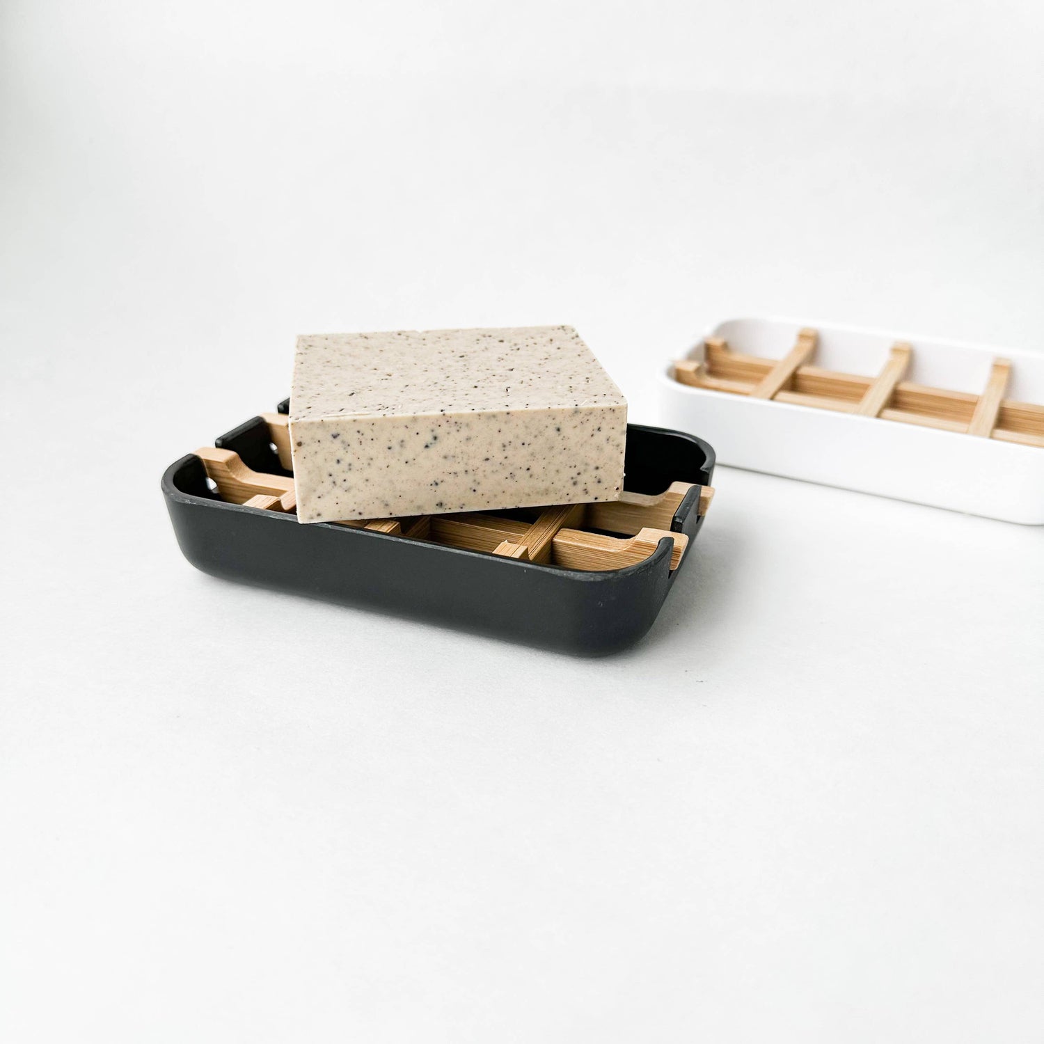 A black biodegradable soap dish with a bar of natural soap on top, and a white dish of the same style but without soap in the background