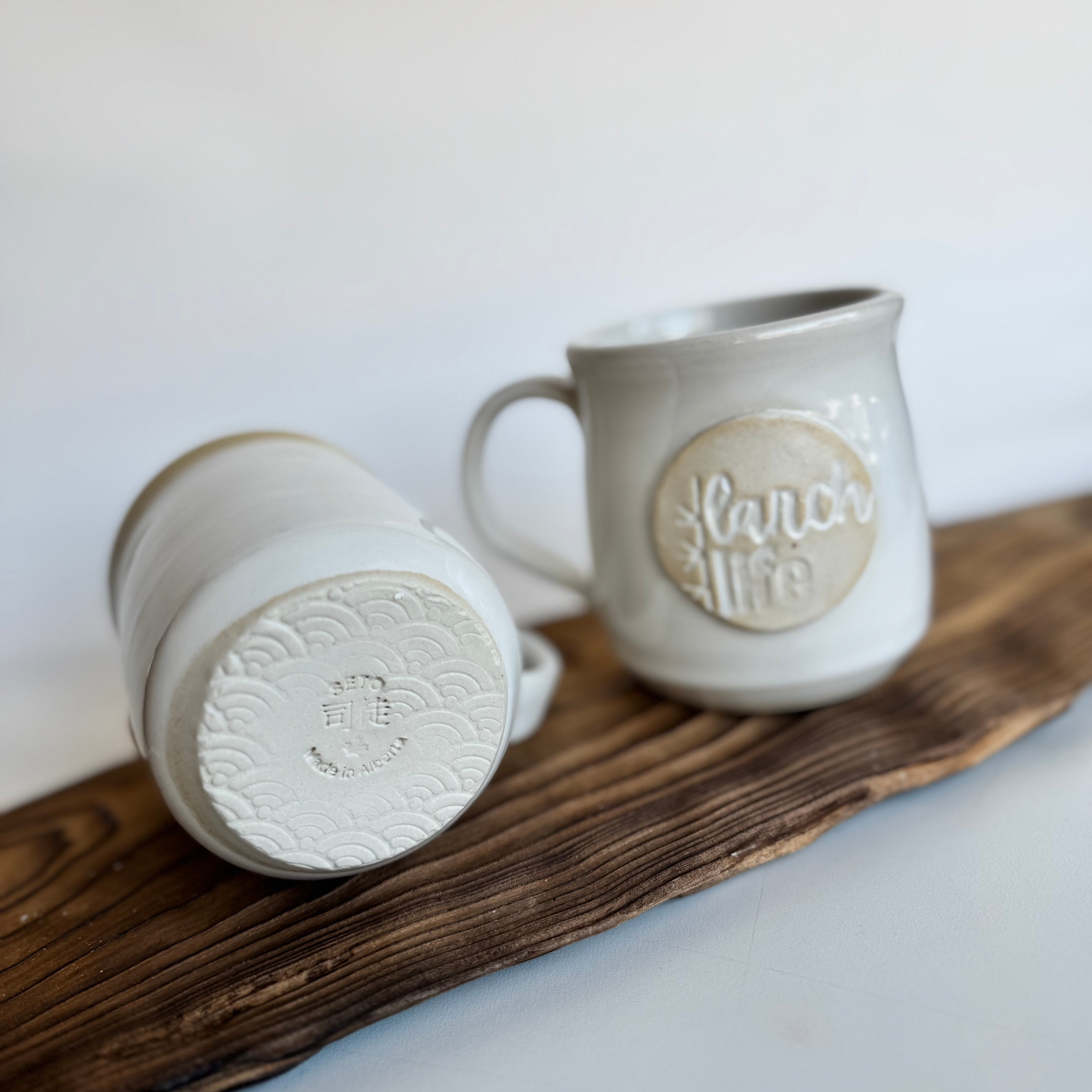 Two handmade mugs by Melba Seto featuring the Larch Life logo, one is turned on its side to show the bottom