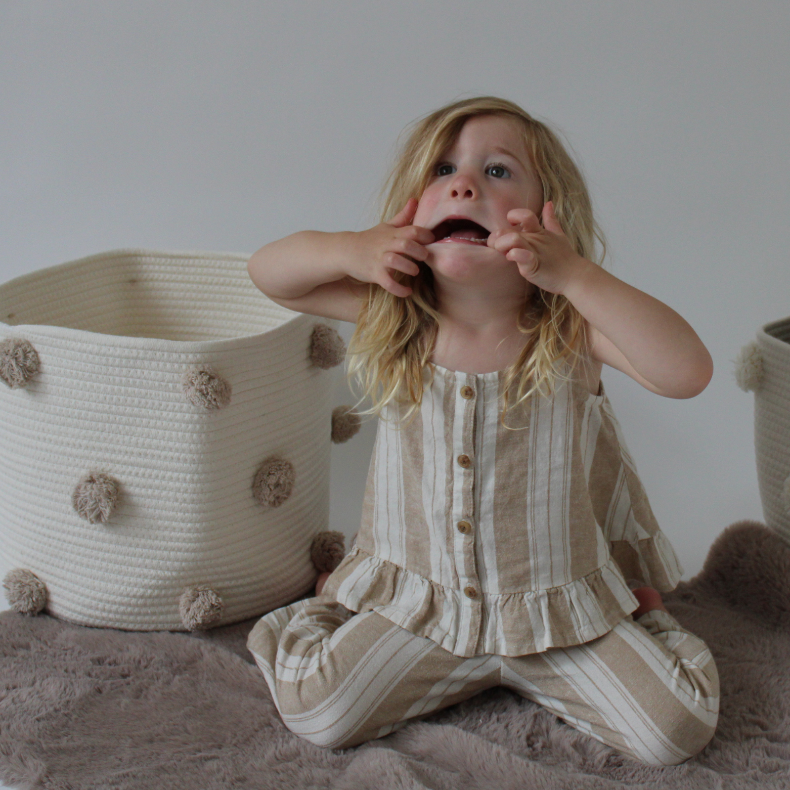 A child plays in front of some handmade natural cotton baskets with taupe pom-poms