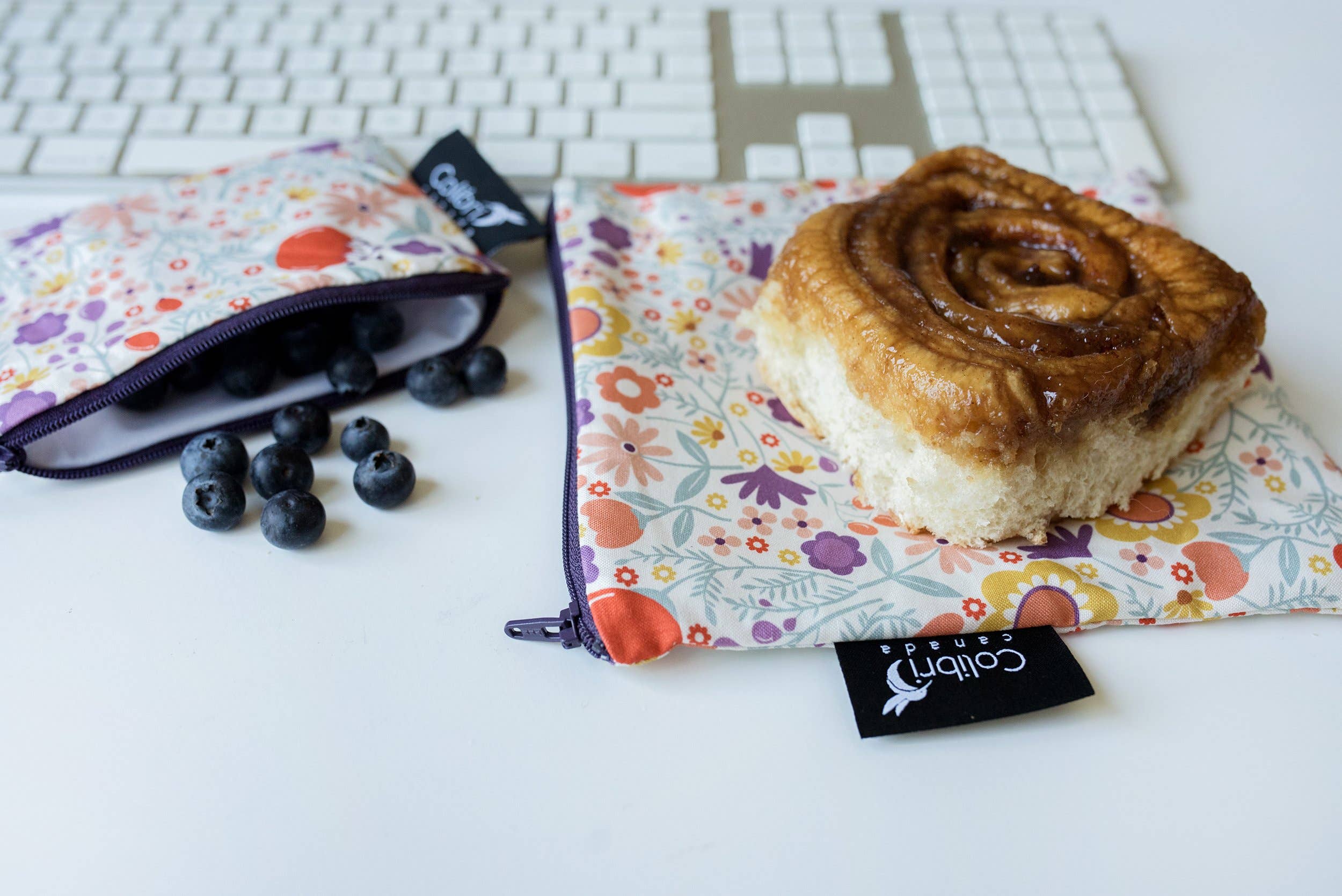 A cinnamon bun sits on top of a flower-motif large zip bag, with a small sized flower-motif bag next to it holding blueberries