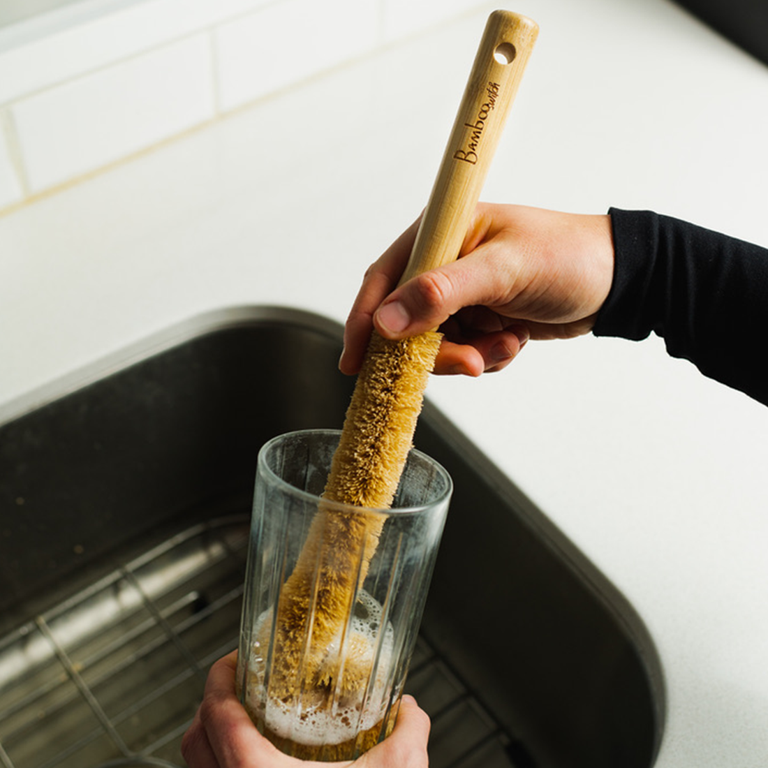 A person using an extra long bamboo and coconut husk bottle brush to clean a glass