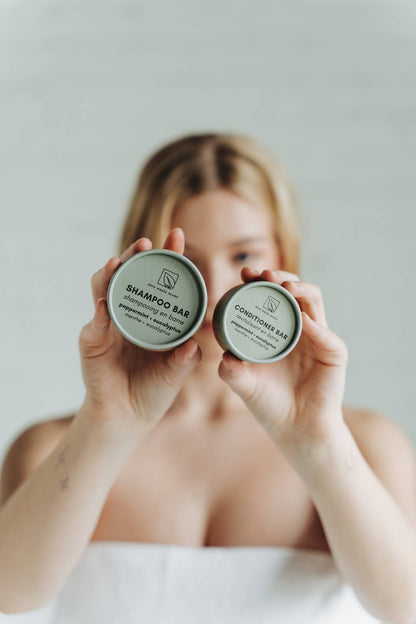 A person holds the packaging for both a sustainable solid shampoo and conditioner bar in front of their face