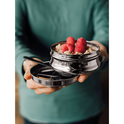 A person holding a small stainless steel snack container with granola and raspberries inside