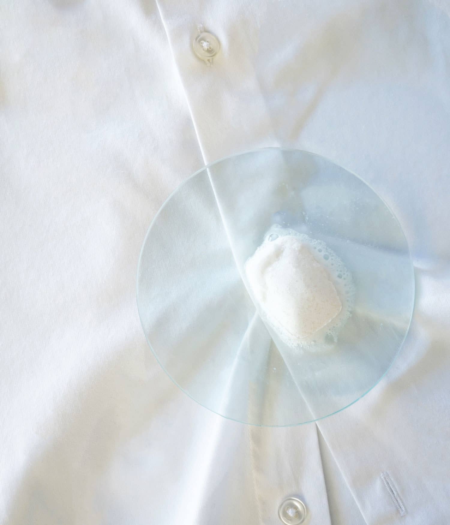 A half-dissolved TANIT laundry detergent tab on a glass dish with a shirt behind